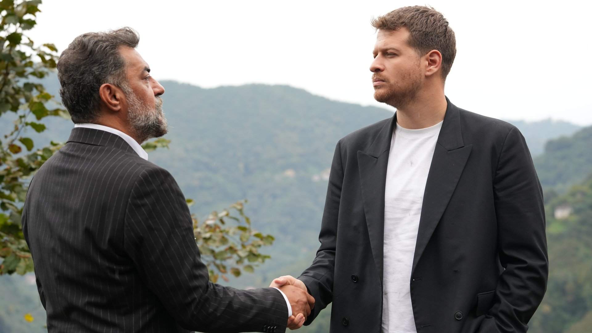 Taşacak Bu Deniz dizisinde yeşil tepelerin önünde tokalaşan iki adam
